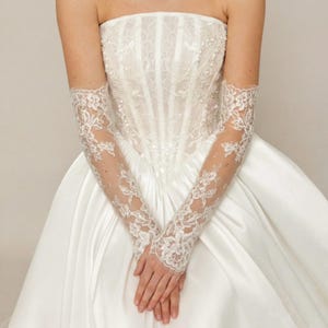 Puede incluir: Vestido de novia blanco sin tirantes con corpi&ntilde;o ajustado y falda amplia. El vestido presenta delicados detalles de encaje y se combina con guantes largos de encaje transparente. Los guantes se extienden desde la mu&ntilde;eca hasta la parte superior del brazo.
