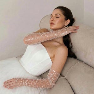 May include: A strapless white wedding dress with a tulle skirt. The bride wears sheer, long gloves adorned with sparkling sequins. The model is wearing a pair of gold earrings with a pearl drop. The bride is reclining on a beige sofa.
