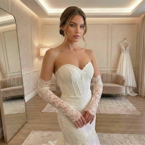 Op de afbeelding: Een ivoorkleurige strapless trouwjurk met een hartvormige halslijn en een nauwsluitend lijfje met gedetailleerd kantwerk. De jurk is gecombineerd met bijpassende transparante kanten mouwen die van de polsen tot de bovenarmen reiken.