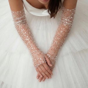 May include: Sheer, long bridal gloves adorned with numerous sequins. The gloves are a light nude color and cover the arms from the fingertips to the upper arm. The background is a white, layered fabric, likely a wedding dress.