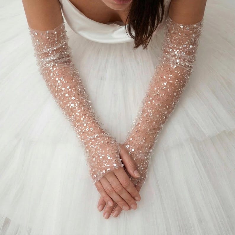 May include: Sheer, long bridal gloves adorned with numerous sequins. The gloves are a light nude color and cover the arms from the fingertips to the upper arm. The background is a white, layered fabric, likely a wedding dress.
