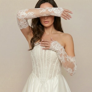 Puede incluir: Un vestido de novia blanco sin tirantes con falda de sat&eacute;n y corpi&ntilde;o detallado. La novia lleva guantes largos de encaje transparente con motivos florales. Los guantes se extienden m&aacute;s all&aacute; de los codos.