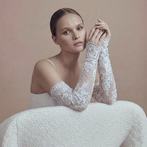 May include: Elegant white lace bridal gloves. The gloves feature intricate floral lace patterns and extend from the wrist to the upper arm. The sheer fabric allows a glimpse of the skin beneath. Perfect for a wedding or special occasion.