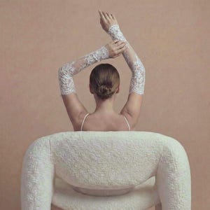 May include: A person wearing white lace arm sleeves, seated in a white, textured chair. The person's arms are raised, and their hair is styled in a bun. The background is a solid, muted peach color.