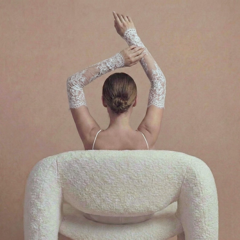 May include: A person wearing white lace arm sleeves, seated in a white, textured chair. The person's arms are raised, and their hair is styled in a bun. The background is a solid, muted peach color.