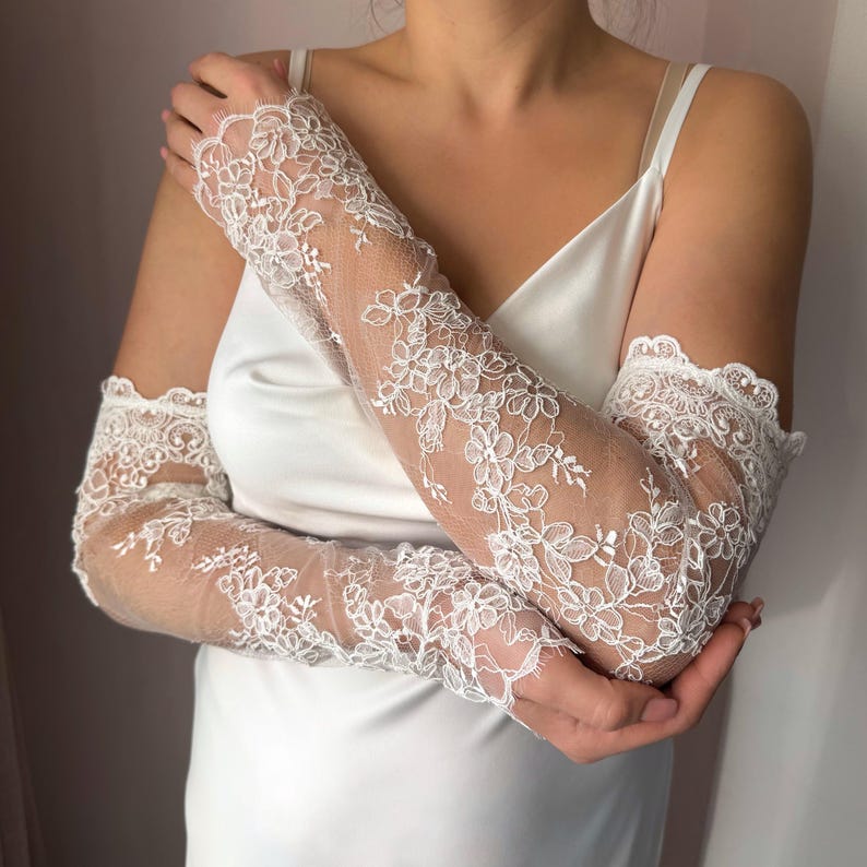 May include: White lace bridal gloves with floral patterns. The gloves are long-sleeved, extending from the fingertips to the upper arm. The lace is detailed with delicate flower designs and scalloped edges.