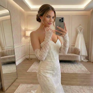 Op de afbeelding: Een strapless witte trouwjurk met een sweetheart halslijn en een zeemeermin silhouet. De jurk heeft ingewikkelde kanten details en wordt gecombineerd met bijpassende kanten handschoenen. Een tweede trouwjurk is zichtbaar op de achtergrond.
