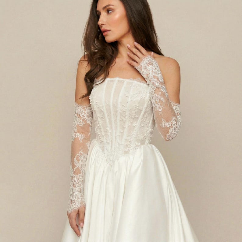 Puede incluir: Vestido de novia blanco sin tirantes con corpi&ntilde;o ajustado y falda amplia. El corpi&ntilde;o est&aacute; adornado con delicados bordados florales. El vestido se combina con guantes largos de encaje transparente, a&ntilde;adiendo un toque de elegancia. El estilo general es cl&aacute;sico y rom&aacute;ntico.
