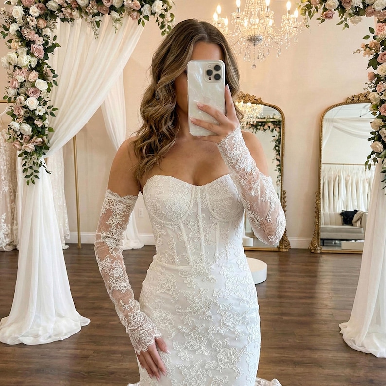 May include: A strapless white wedding dress with intricate lace detailing. The dress features a fitted bodice and a flowing skirt. Matching lace sleeves are worn. The background includes floral arrangements, mirrors, and a chandelier.