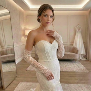 Op de afbeelding: Een strapless ivoorkleurige trouwjurk met een korset-stijl lijfje met gedetailleerd kant en bijpassende lange, doorschijnende kanten mouwen die tot aan de polsen reiken. De jurk heeft een nauwsluitend silhouet dat onderaan uitloopt.