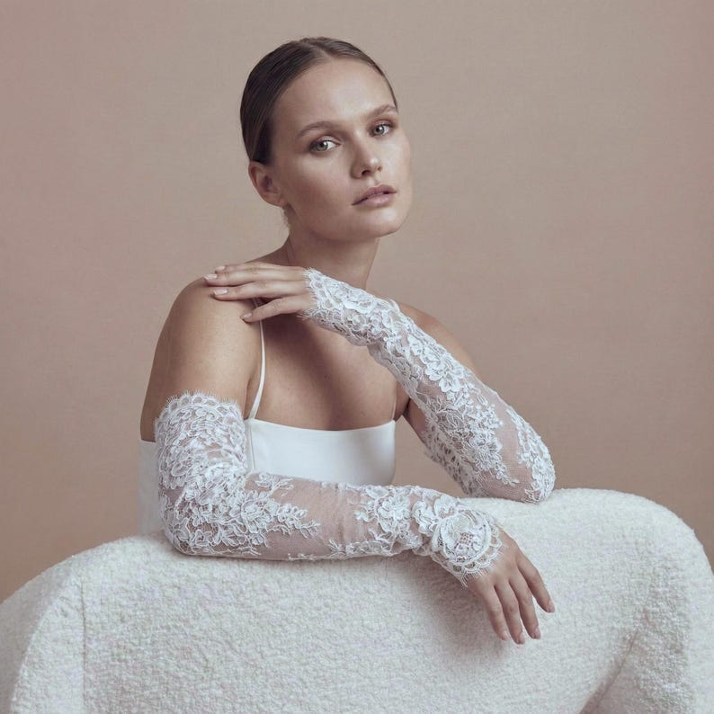 May include: White lace bridal gloves with intricate floral patterns. The gloves are long, covering the arms up to the upper arm. The delicate lace design adds a touch of elegance to the bridal wear.