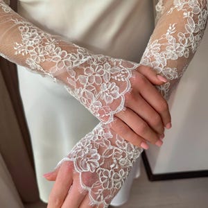 May include: Delicate white lace sleeves with floral patterns. The sleeves are sheer, revealing the skin beneath, and are likely part of a bridal or formal wear ensemble. The lace features intricate detailing and scalloped edges.