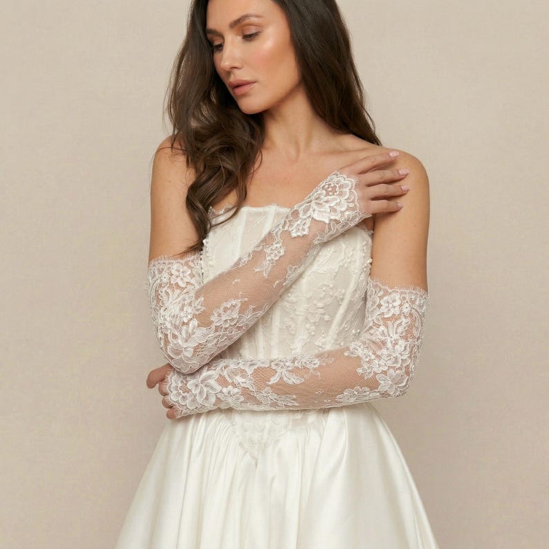 Puede incluir: Una novia con un vestido de novia blanco y largos guantes de encaje transparente. Los guantes presentan un delicado estampado floral y se extienden desde la punta de los dedos hasta la parte superior del brazo. El vestido es sin tirantes y tiene un corpi&ntilde;o ajustado.