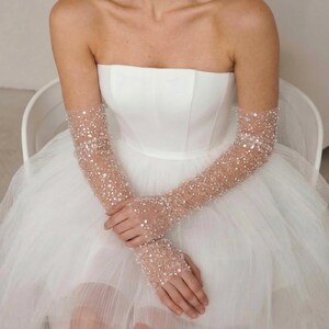 May include: A strapless white wedding dress with a full tulle skirt. The sleeves are sheer, covered in small, sparkling embellishments. The sleeves extend to the fingertips. The person is seated in a white chair.
