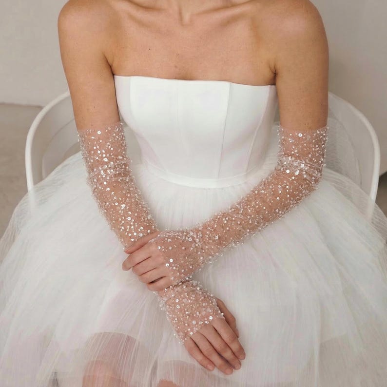 May include: A strapless white wedding dress with a full tulle skirt. The sleeves are sheer, covered in small, sparkling embellishments. The sleeves extend to the fingertips. The person is seated in a white chair.