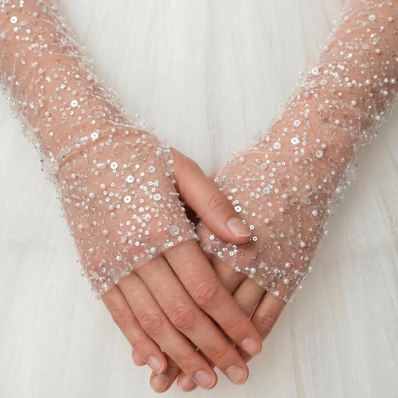 May include: Close-up of sheer, light pink gloves adorned with sequins and small pearls. The gloves cover the hands and extend up the forearms. The background is a soft, white fabric, creating a delicate and elegant aesthetic. Ideal for weddings or special occasions.