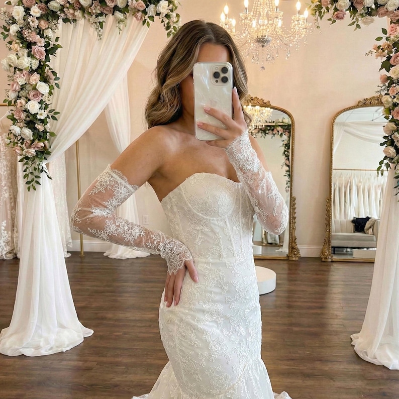 May include: A strapless white wedding dress with intricate lace detailing. The dress features a fitted bodice and a flared skirt. The bride is wearing sheer lace sleeves. The background includes floral decorations and mirrors.
