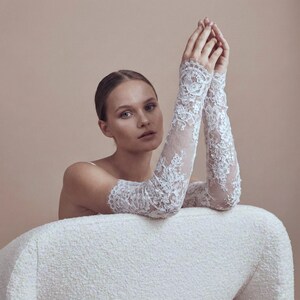 May include: Elegant white lace gloves with intricate floral patterns. The gloves extend to the upper arm, creating a sophisticated look. The model is seated on a white textured chair, with arms raised. The background is a neutral beige.