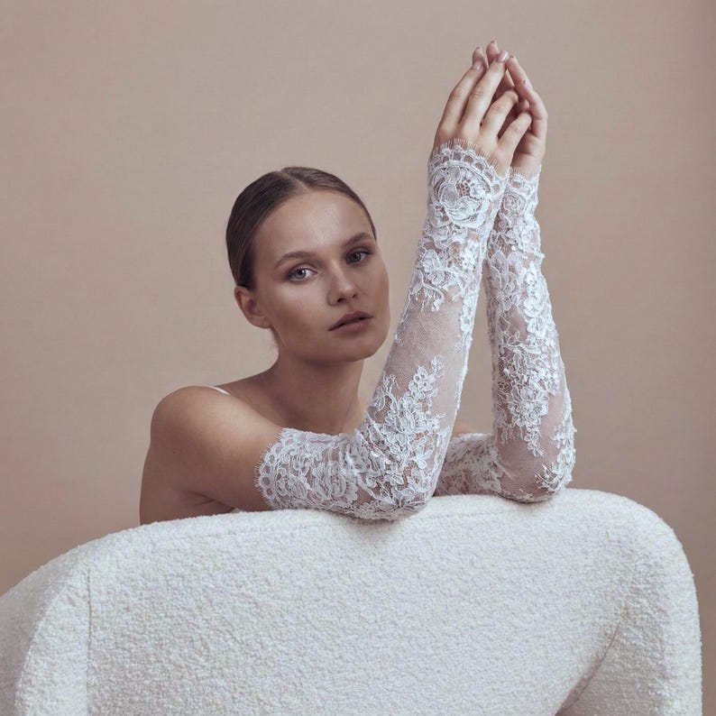 May include: Elegant white lace gloves with intricate floral patterns. The gloves extend to the upper arm, creating a sophisticated look. The model is seated on a white textured chair, with arms raised. The background is a neutral beige.