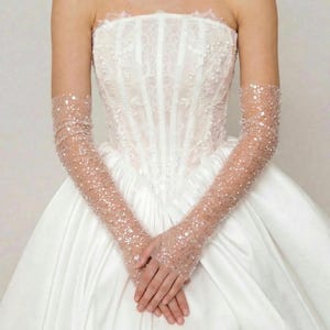 May include: A white strapless wedding dress with a satin skirt and a detailed bodice. The dress is paired with sheer, elbow-length gloves adorned with sparkling sequins. The gloves add a touch of glamour to the bridal ensemble.
