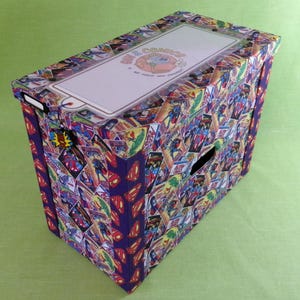 Może przedstawiać: Prostokątne pudełko do przechowywania pokryte panelami komiksowymi z Supermanem i innymi postaciami. Pudełko ma białą pokrywę z napisem "Duff & Coasters". Pudełko ma wycięcie na uchwyt z boku.