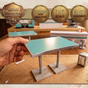 Puede incluir: Una mesa de comedor en miniatura con una tapa turquesa y borde plateado, sostenida por dos patas plateadas. La mesa es parte de un taller de manualidades de papel, con el texto "Retro Miniatures" y "Easy Assembly" visible.