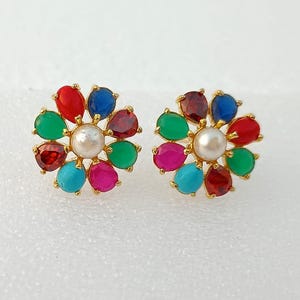 May include: A pair of gold-toned stud earrings. Each earring features a flower design with colorful gemstones in red, blue, green, pink, and turquoise, surrounding a central pearl. The earrings are set against a white background.