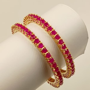 May include: Two gold-toned bangles featuring a continuous row of round, vibrant pink stones set in a metallic frame. The circular bracelets have a polished finish and a consistent design, showcasing a bright, saturated color palette throughout.