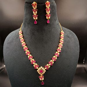 May include: A gold-toned necklace and earring set. The necklace and earrings feature a repeating pattern of pink and white stones. The necklace has a central floral pendant with a teardrop-shaped pink stone. The earrings have a teardrop-shaped pink stone.