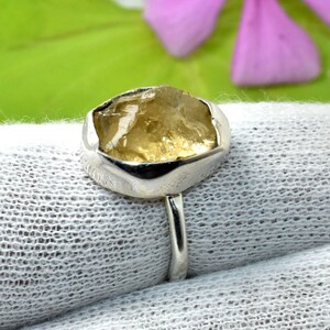 Puede incluir: Un anillo con una piedra preciosa amarilla, cruda y translúcida, engastada en un bisel de metal pulido de color plateado. La piedra tiene una superficie irregular y facetada con inclusiones naturales, y la banda es de metal liso.
