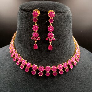 Pode incluir: Um colar dourado e um conjunto de brincos a condizer. As joias apresentam um design floral com pedras preciosas rosa vibrantes. O colar é exibido num suporte preto, mostrando os detalhes intrincados do colar e dos brincos.