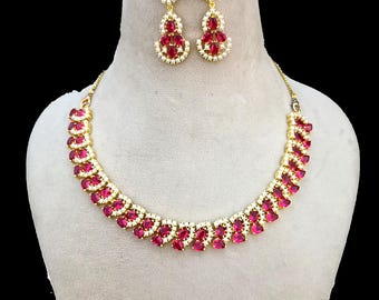 May include: A gold-toned necklace and earring set. The necklace and earrings feature oval-shaped, ruby-red gemstones and small, clear stones. The jewelry is displayed on a mannequin against a black background. The earrings are drop style.