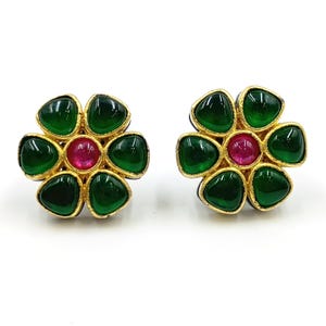 May include: A pair of flower-shaped stud earrings featuring a central round pink stone surrounded by six teardrop-shaped green stones. The stones are set in a textured gold-toned metal frame, creating a symmetrical floral design for each earring.