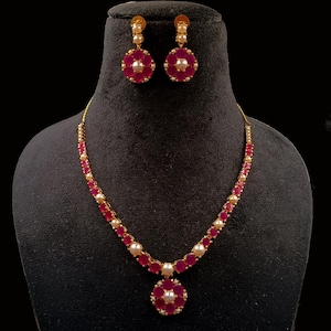 May include: A gold-toned jewelry set featuring a necklace and matching earrings. The necklace and earrings have a floral design with ruby-red stones and pearl accents. The set is displayed on a black mannequin head against a black background.