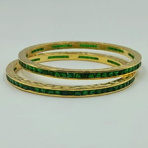 May include: Two gold-toned bangles featuring a continuous channel setting of square-cut, vibrant green stones. The metal surface of the bangles has a subtle hammered texture, providing a contrast to the uniform, faceted appearance of the green stones.