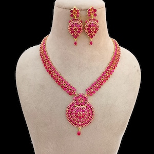 Puede incluir: Un conjunto de collar y pendientes de color dorado con piedras preciosas rosas vibrantes. El collar tiene un colgante grande y circular y una cadena delicada. Los pendientes son redondos con un diseño de piedras preciosas a juego. La joyería se exhibe en un maniquí.