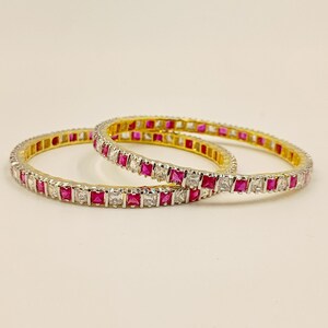 Pode incluir: Dois braceletes circulares com um padrão repetitivo de pedras quadradas alternadas em rosa e branco transparente, fixadas numa estrutura metálica. O design consiste numa fila contínua destas pedras facetadas dispostas lado a lado.