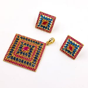 Puede incluir: Un conjunto de joyería dorada que consta de un colgante cuadrado y dos pendientes cuadrados a juego. Cada pieza presenta una piedra roja ovalada central rodeada de piedras azules talla marquesa y un borde de pequeñas piedras rosas.