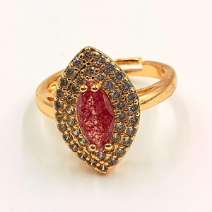 Könnte beinhalten: Ein goldfarbener Ring mit einem zentralen, marquiseförmigen roten Stein, der von einem Kranz aus kleinen, klaren, funkelnden Steinen umgeben ist. Die Ringschiene ist verstellbar und hinten offen, mit einer polierten Oberfläche und Fassung.