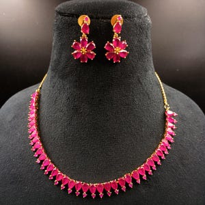 May include: A gold-toned necklace and earring set. The necklace features a series of teardrop-shaped, magenta-colored stones. The earrings are flower-shaped with magenta stones. The jewelry is displayed on a black mannequin.