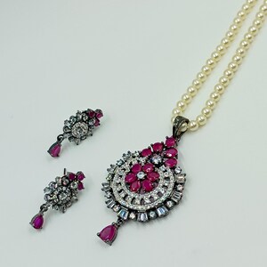May include: A jewelry set consisting of a necklace and two earrings. The necklace features a string of cream-colored pearls and a pendant with dark metal, clear crystals, and deep pink teardrop and petal-shaped stones. Matching earrings.