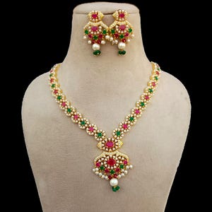 Peut inclure: Un ensemble collier et boucles d'oreilles doré. Le collier présente un pendentif avec des pierres précieuses rouges et vertes, entouré de perles blanches. Les boucles d'oreilles reprennent le design du collier. L'ensemble est présenté sur un mannequin.