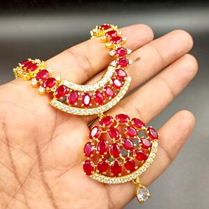 Puede incluir: Collar dorado con diseño de media luna, adornado con piedras rojas de corte ovalado y pequeños cristales transparentes. El collar presenta un colgante con un grupo de piedras rojas y un cristal en forma de lágrima.
