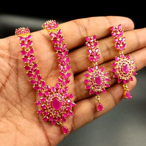 Puede incluir: Un conjunto de joyas de tono dorado con un collar y pendientes. El collar y los pendientes están adornados con vibrantes piedras preciosas rosas. El collar tiene un colgante floral con una gema en forma de lágrima.