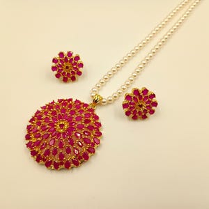May include: A jewelry set consisting of a necklace and two matching earrings. The necklace features a string of white pearls with a large, circular gold pendant set with numerous teardrop-shaped fuchsia stones. The earrings match the pendant.