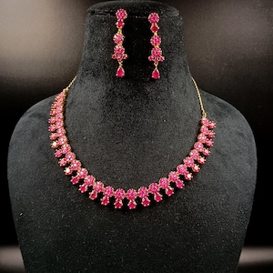May include: A gold-toned necklace and matching earrings set. The jewelry features a repeating floral design with vibrant pink gemstones. The necklace and earrings are displayed on a black mannequin head against a dark background.