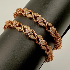 May include: Two rose gold-toned bangles featuring a repeating twisted vine pattern. Each bangle is adorned with small, clear, sparkling stones set into floral motifs and leaf-shaped segments that wrap around the entire circumference of the band.