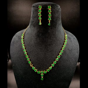 May include: A gold-toned necklace and matching earrings set. The jewelry features a series of vibrant green gemstones. The necklace has a delicate design, with the gemstones set closely together. The earrings are long and dangle, with a similar gemstone arrangement.
