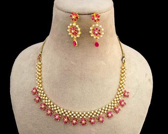 May include: A gold-toned necklace and earring set. The necklace features a floral design with pink gemstones and white pearls. The earrings have a similar floral design with a teardrop-shaped pink gemstone. The jewelry is displayed on a mannequin.