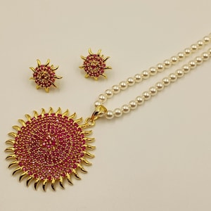 Puede incluir: Un conjunto de joyería compuesto por un collar y dos pendientes. El collar presenta una hilera de perlas blancas y un gran colgante circular de tono dorado incrustado con pequeñas piedras de color rosa intenso en forma de sol.